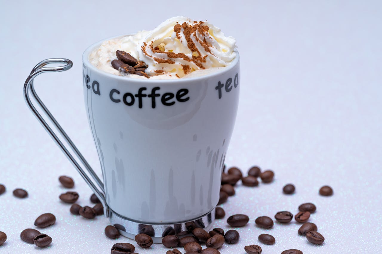 Creamy coffee topped with whipped cream and cinnamon, surrounded by coffee beans on a white background.
